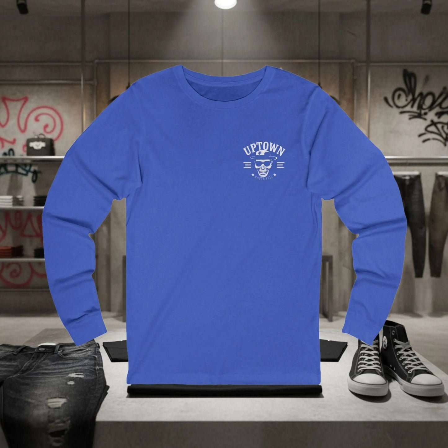 Blue long-sleeve shirt with 'Uptown' logo on a stand in a room with shoes and graffiti.